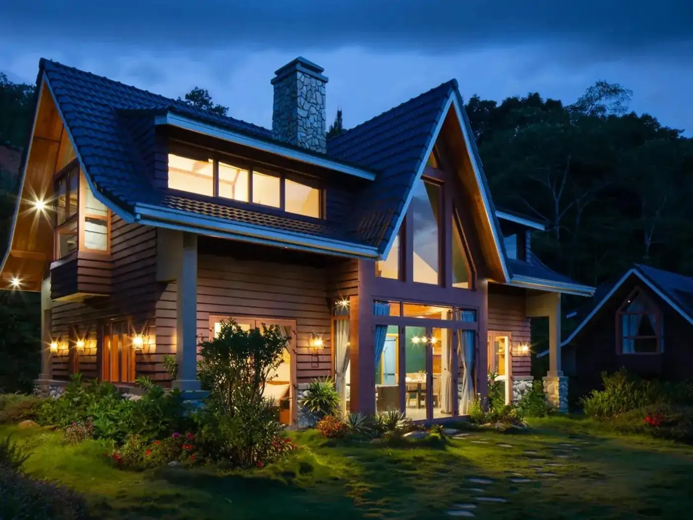 A two-storey timber house at dusk with lit windows and a steeply pitched roof